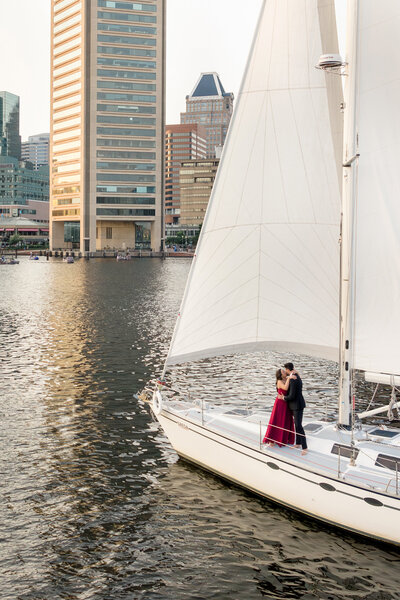 A couple embraces on a sailboat wearing a black suit and a red gown. The boat is in a calm harbor with office buildings in the background, reflecting cityscape and water under soft sunlight.