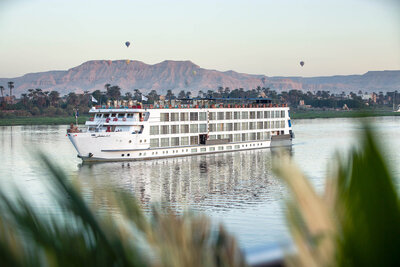 A large white river cruise ship sails on a calm river, with palm trees along the shore and desert mountains in the background. A few hot-air balloons float in the sky above the landscape.