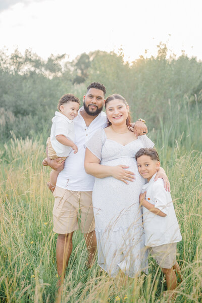 A beautiful family of four, soon to be a family of five, hugging each other in a large open field, sharing a joyful and heartfelt moment together