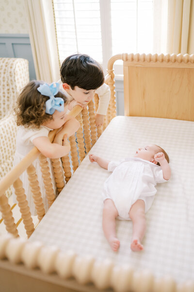 Big brother and sister lean over crib railing to look at newborn baby brother during photos