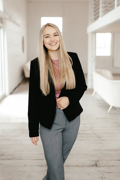 In a bright, modern room, a woman with long blonde hair smiles as she walks. Wearing a black blazer over a pink top and gray pants, she embodies the elegance captured by Amber Bauer Photography. The light wooden floors and beige couch enhance the room's welcoming ambiance.