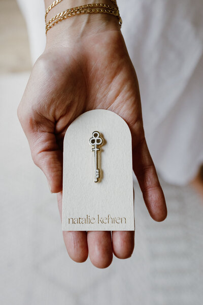 Hand holding a beige card with a gold key pin and the name 'natalie kehren' embossed on it.