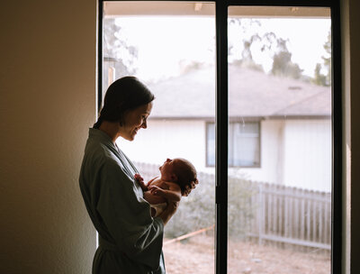 A tender moment in the newborn stage where a mother  looks down at her newborn baby as the baby looks up into her eyes standing in front of the window looking out where her other child is playing.