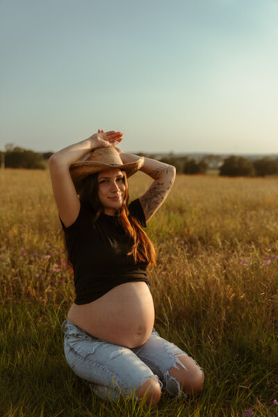 Pregnant woman in a cowgirl hat kneeling in a golden field at sunset, holding her belly and smiling during a rustic maternity photoshoot.