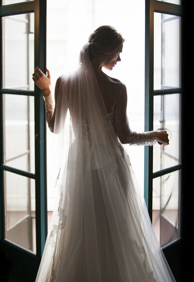 Kohnur bride in a long sleeve lace gown and simple veil silhouette against the French doors of a classic estate.