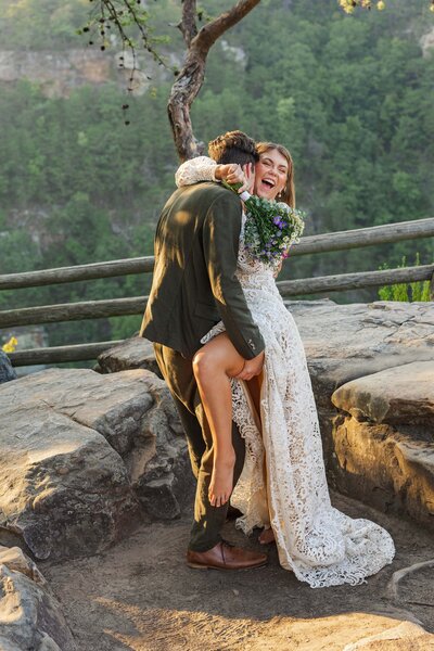 A bride and groom share a sweet embrace after eloping. The groom has one arm around the brides waist while the other arm holds her thigh up so it wraps around him. The bride has her arms around the grooms neck and is laughing.