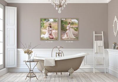 A bathroom with a taupe clawfoot bathtub with a shower head on a hose and light grey wooden floors and a light brown end table holding a white vase with ivory flowers and two pictures on the wall of a pregnant woman in a white lace dress and a floral crown on her head hanging on the wall and a crystal chandelier hanging in the middle of the ceiling right above the bathtub.