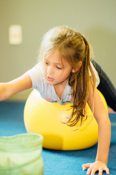 elementary aged girl laying on her belling on a yoga ball while she balances to throw bean bags in a container