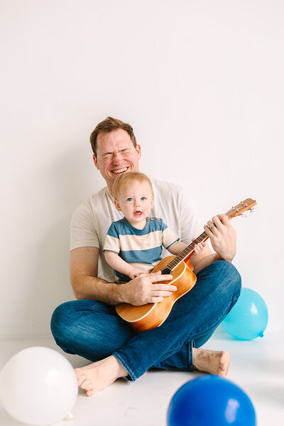 Dad with baby son in his lap holding a guitar with blue balloons around them