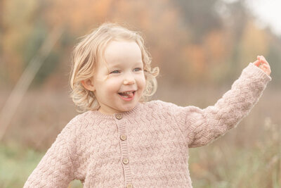 Toddler girl in light pink sweater with golden curls is smiling off camera with her tongue out a little bit and there are beautiful fall leaves in the background behind her. 