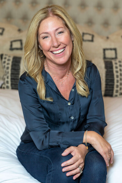A woman with long blonde hair, wearing a dark blue blouse and jeans, sits on a bed, smiling at the camera with her arms resting on her knees—the perfect image of a web design client for Sweet Pea Ink Creative.