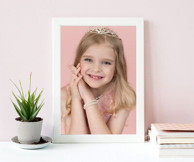 Framed photo of a smiling young girl with a tiara and pearl bracelet against a pink background. She's propped on her hands. Nearby, a small potted plant and stacked notebooks add a cozy touch.