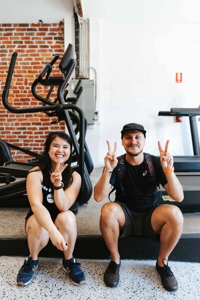 2 personal trainers sitting on a treadmill smiling in a 24 hours dog friendly gym.