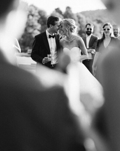 Couple de mariés rient ensemble, front à front, verre à la main, durant un discours lors du cocktail. Image en noir et blanc.