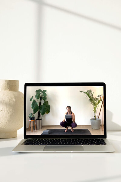 Open laptop on a white surface showing a video of Alice, sitting on a mat in a squat position holding a tablet, with indoor plants in the background, depicting a calm teaching environment for an online yoga course.