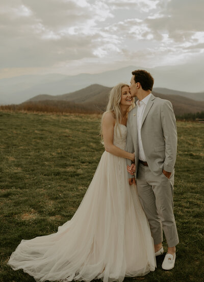 photo of couple embracing on top of Max Patch Mountain in North Carolina, near the Tennessee border. In the background the mountains are surrounding them. The woman is wearing a long flowing white dress, he is wearing a grey suit blazer and white dress shirt.