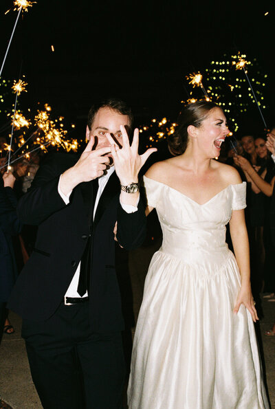 Bride and groom walk out of their reception into a sparkler exit while bride laughs and groom points to his ring