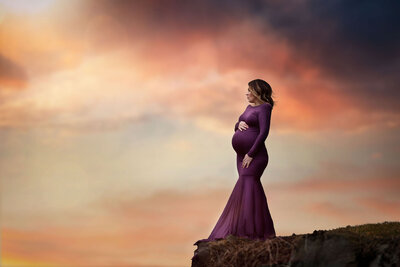 pregnant woman wearing a plum gown standing on a cliff with a dramatic sunset in the background