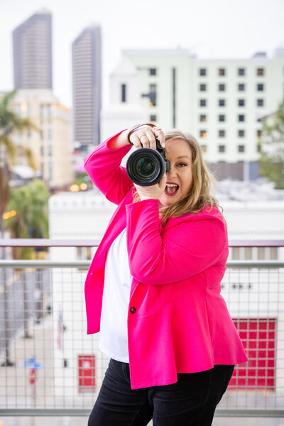 Megan Sethia, Seattle brand photographer and founder of She Plays with Fire, playfully posing with her camera in a bold pink blazer on a city rooftop—capturing vibrant, confident brand imagery for creative entrepreneurs.