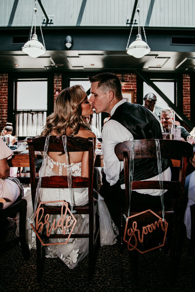 Bride and groom sharing a kiss while seated at their sweetheart table with “bride” and “groom” chair signs during their San Francisco elopement reception, captured by San Francisco elopement photographer Kristin Smith.