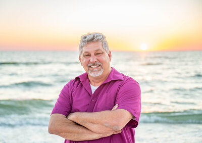 Photo of John Corbellini smiling in a  pink collared shirt standing in front of a beach sunset