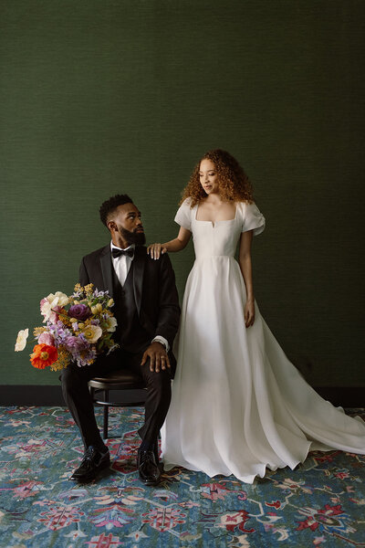 Editorial wedding portrait of a bride and groom in a modern indoor setting, featuring the groom seated in a black tuxedo holding a colorful garden-style bouquet with poppies, ranunculus, peonies, and roses, while the bride in a minimalist square-neck white wedding gown with puff sleeves stands beside him against a deep green wall. Romantic, contemporary wedding inspiration with bold floral design and elegant fashion styling.