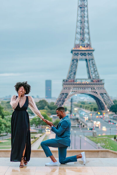 Black couple during surprise proposal in Paris — woman shielding her eyes as the man waits to propose with the Eiffel Tower in the background.