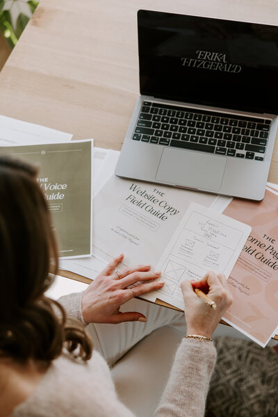The Brand Voice Field Guide and stack of copywriting papers on a wooden desk with colorful pens, copywriting books, and AirPods