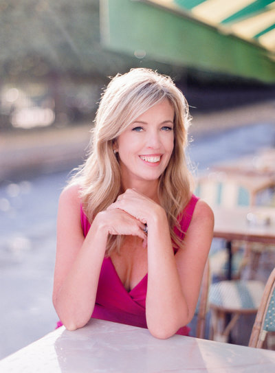 Photographer Cari Long smiles while setting at a cafe table outside in Paris wearing a pink dress