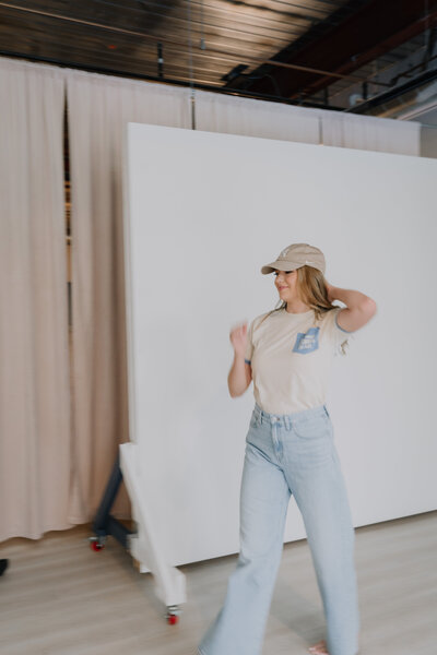 Megan in jeans and a hat standing in front of a studio backdrop