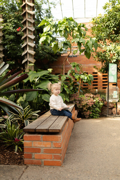 toddler girl sits on brick bench in a tropical conservatory holding a book looking off into the distance