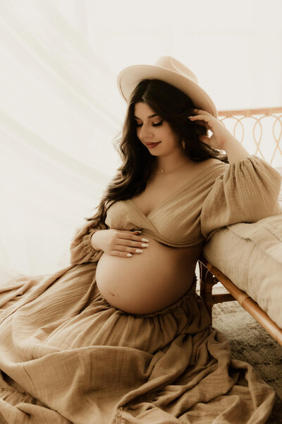 Expecting mother looks at her baby bump while wearing a hat and beige skit with a matching top.