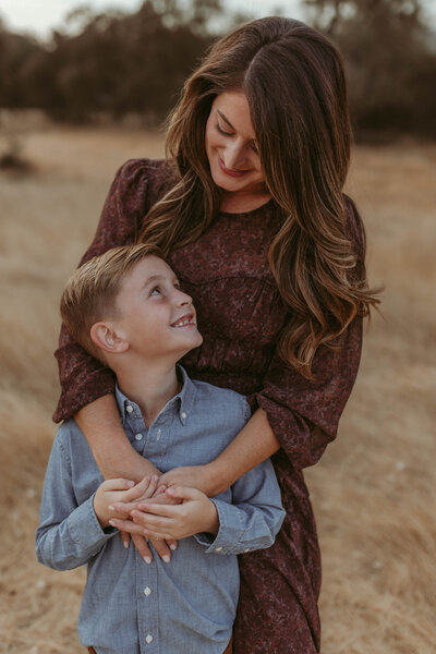 A mother holds her young son and looks lovingly into his eyes while he gives her a toothy grin back. 
