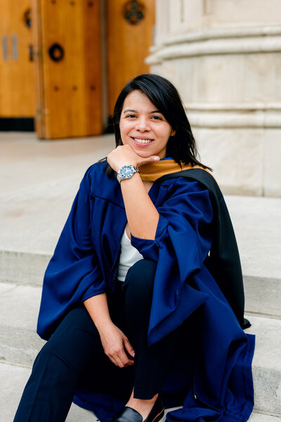 College graduate wearing academic hood and gown while seated on stone steps.
