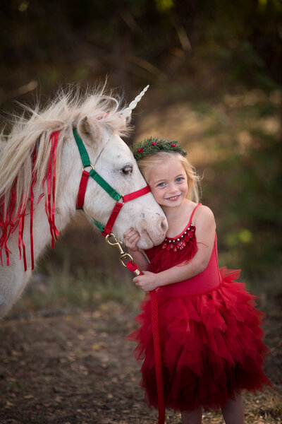 girl-holding-unicorn-rein-in-holiday-red-dress-and-unicorn-has-holiday-ribbons-inarlington-tx