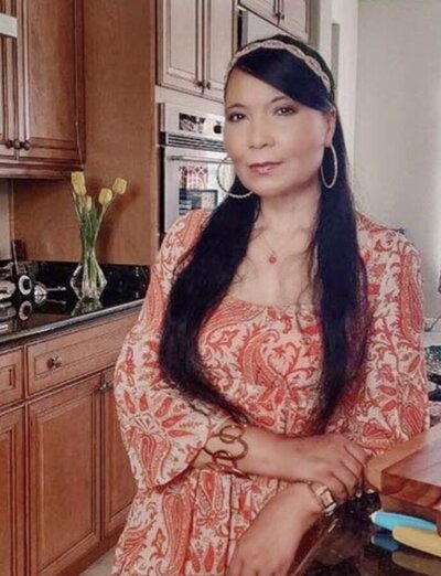 Image: photo of Regina. Standing in her kitchen wearing an orange and beige floral print blouse. hair over both shoulders. yellow tulips in a glass vase in the background.