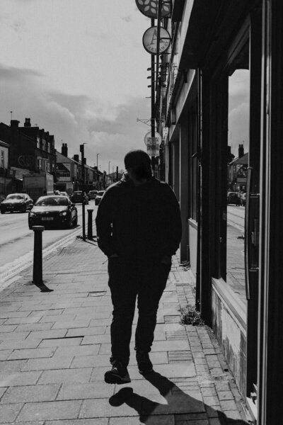 Black and white photo of a person walking down a city sidewalk, with cars and buildings lining the street. The person is silhouetted, creating a dramatic contrast against the urban backdrop.