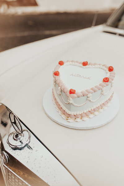 pink and white heart shaped cake with cherries on top sitting on top of a vintage car