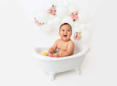 Brooklyn, NY Cake Smash Photographer captures baby girl getting messy with her arms covered in cake and eating a handful of icing. Baby is wearing a pink floral crown and fresh flowers top the white cake.