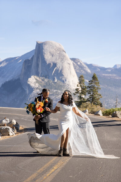 A bride in hiking boots walks with her groom during their Yosemite elopement at Glacier Point, with Half Dome towering in the background. Captured by Yosemite elopement photographers, this moment blends adventure with romance in an iconic national park setting.