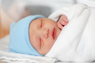 Close-up of swaddled newborn’s face and hand by his chin, wearing a blue hat