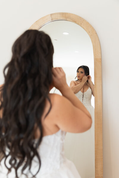 The Lakehouse Inn MA  | Kelsey Sheehan Photography Timeless Rhode Island Weddings | A bride in a white gown adjusts her earrings in front of a tall mirror. Her long, dark hair cascades down her back. The room is softly lit, conveying calmness.