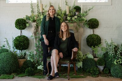 Jazmin stands next to Alex, who is sitting in a chair legs crossed. the Luna & Co Owners look happy and like they make a great team. They're in front of a mantel decorated with an abundance of greenery and floral arrangements.