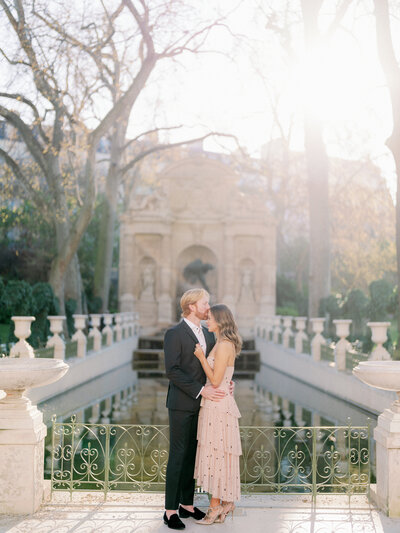 a man in a suit facing a woman in a pale pink tulle dress kissing her forehead in front of a pond at jardin du luxmebourg and and embracing with the sun shining behind them