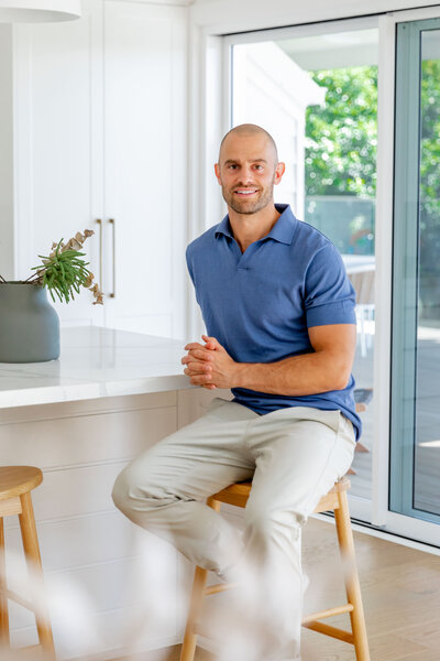 Jon Moses wearing a blue polo shirt, beige pants sitting on a wooden stool