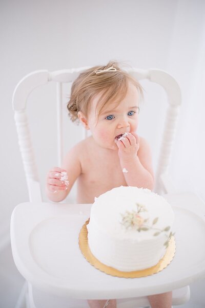 Baby girl sitting in white high chair eating cake for 1st birthday cake smash at stamford ct portrait studio