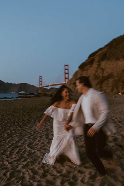 Bride and groom running down San Francisco beach with Golden Gate bridge behind them