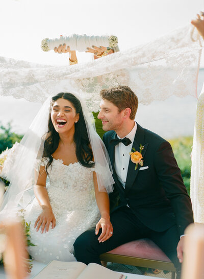 A bride and groom are seated outdoors during a wedding ceremony, smiling as a lace veil is held above them.