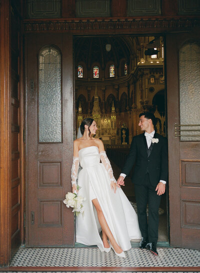 Person in a black suit admiring their parnter in a white wedding dress leaning against the wall at City Hall