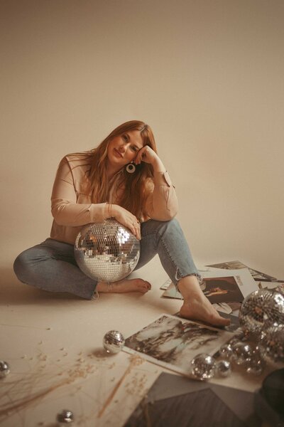 Gillian Oler Photographer brand photoshoot. She is wearing blue jeans and a tan shirt, sits in a photoshoot room, cradling a disco ball in her arms. There are taylor swift vinyl records and flowers on the floor.